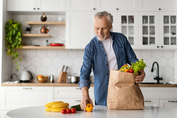 Handsome Senior Gentleman Unpacking Paper Paper Bag With Groceries In Kitchen Interior At Home, Happy Elderly Male Putting Fresh Fruits And Vegetables On Table After Successful Food Shopping - Photo, Image
