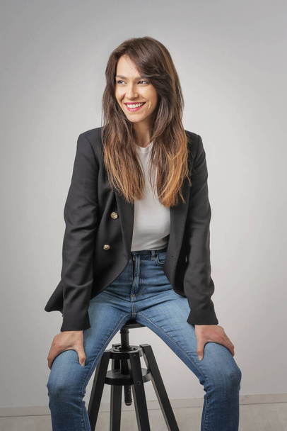 Studio portrait of attractive woman wearing blazer and laughing while sitting at isolated grey background. Copy space.  - Φωτογραφία, εικόνα
