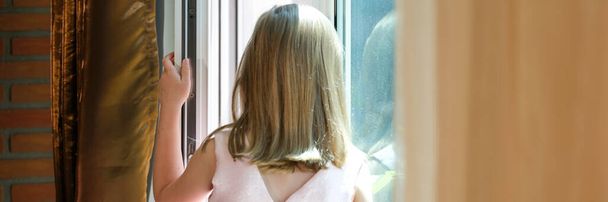 Close-up of girl in pink dress standing on windowsill at open window. Danger of falling from height concept - Foto, afbeelding
