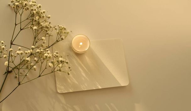 Burning candle, flower twig and empty paper card. Evening Light and shadows minimalism still life template background. Top view. - Photo, Image
