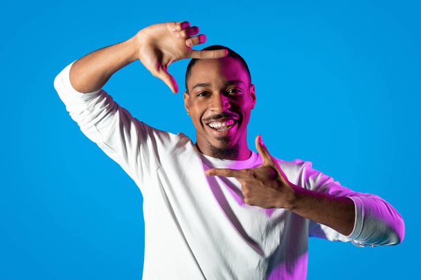 Positiv fröhlich gut aussehender bärtiger junger afrikanisch-amerikanischer Typ in weißem Hemd, mit Fingern Kamera-Geste um das Gesicht, blauer Hintergrund im hellen Licht, Kopierraum - Foto, Bild