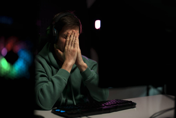Joven jugador de ciberdeporte masculino decepcionado sentado en la mesa con las manos de oración mientras juega videojuego en el ordenador - Foto, imagen