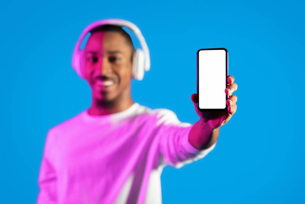 Smartphone mit weißem leeren Bildschirm in afrikanisch-amerikanischer Hand, fröhlicher junger schwarzer Mann mit drahtlosem Kopfhörer, der Handy zeigt, Empfehlung für schöne musikalische Handy-App, blauer Hintergrund, Attrappe - Foto, Bild