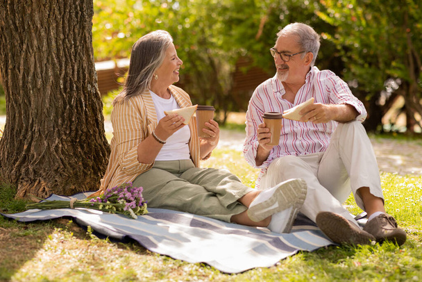 Heureux couple d'âge mûr européen avec tasse de café à emporter, sandwich assis sur plaid, profiter de la date, pique-nique dans le parc le week-end d'été, en plein air. Temps libre, amour, romance, déjeuner et nourriture - Photo, image