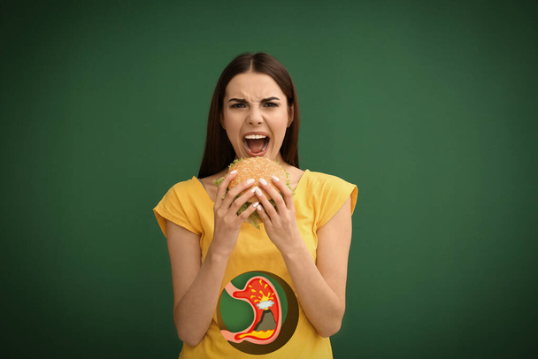 Improper nutrition can lead to heartburn or other gastrointestinal problems. Woman eating burger on green background. Illustration of stomach with erupting volcano as acid indigestion - 写真・画像