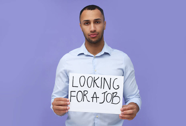 Young unemployed man holding sign with phrase Looking For A Job on violet background - Fotoğraf, Görsel