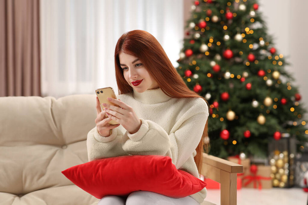 Beautiful young woman using smartphone on sofa near Christmas tree at home - Fotoğraf, Görsel