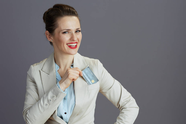 happy elegant 40 years old business woman in a light business suit with credit card against grey background. - Φωτογραφία, εικόνα