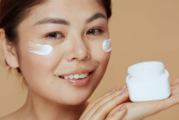 Portrait of modern asian female with facial cream jar and facial cream on face on beige background. - Φωτογραφία, εικόνα