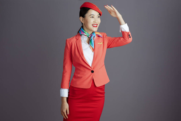 happy modern asian female flight attendant in red skirt, jacket and hat uniform looking into the distance isolated on gray. - Φωτογραφία, εικόνα