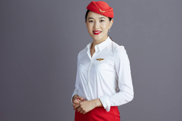 smiling stylish asian female stewardess in red skirt and hat uniform isolated on grey background. - Фото, изображение