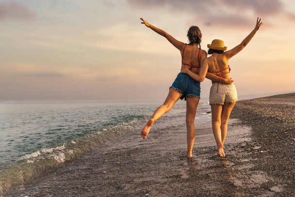 Dva mladí přátelé, jeden se slamákem, se procházejí po pláži při západu slunce. Pozvedají ruce v radostných gestech, vyjadřují lásku a přátelství. Pojmy: přátelství, dovolená, touha po toulkách. - Fotografie, Obrázek