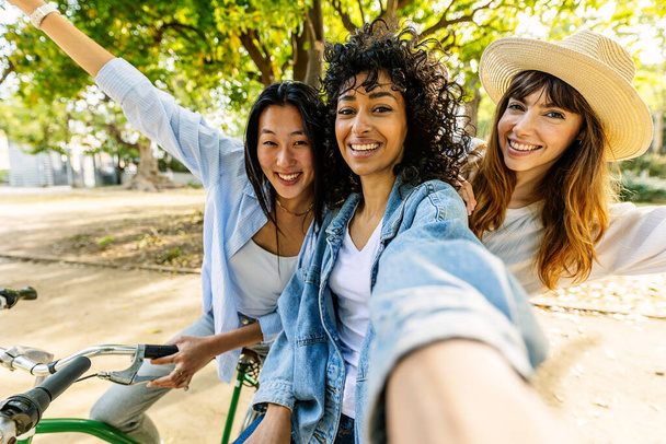 Three happy diverse girls having fun enjoying bicycle ride on city park during summer vacation. Millennial female friends taking selfie portrait together enjoying day off at weekend. - Foto, immagini
