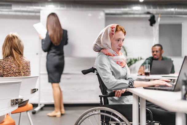 Jeune fille en fauteuil roulant portant un hijab travaillant sur son ordinateur portable au bureau - Photo, image
