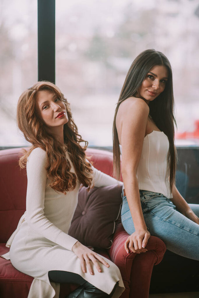 Nahaufnahme von zwei schönen Models mit langen, glatten und lockigen Haaren, die auf dem rosa Sofa posieren - Foto, Bild
