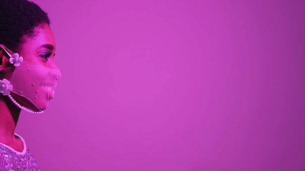 Quarantine fashion. Face accessory. Pandemic party. Purple pink color neon light profile portrait of confident smiling woman in mesh mask on fluorescent empty space background. - Foto, Imagem