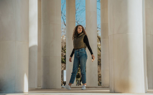 Stunning curly haired girl standing between white pillars and looking away. She is holding a phone and sunglasses in her hand. Full length photo - Foto, Bild