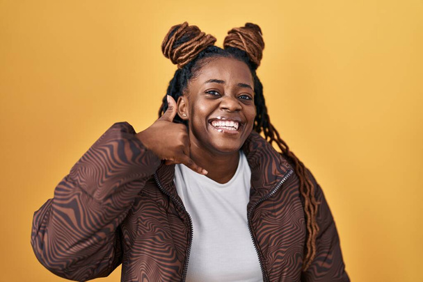 Africká žena s vlasy spletenými do copu, stojící nad žlutým pozadím a usmívající se, mávajíc rukou a prsty, jako by mluvila do telefonu. komunikace konceptů.  - Fotografie, Obrázek