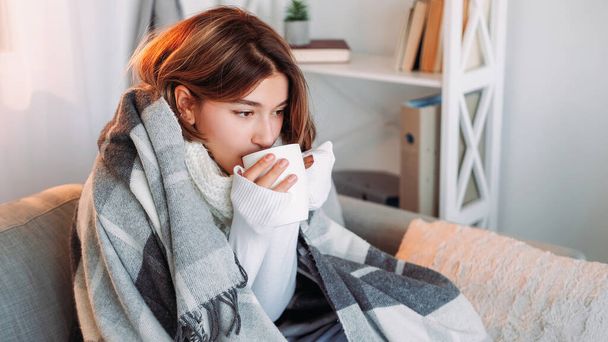 Tea warm. Rest drinking. Autumn shivering. Cold brunette woman in blanket enjoying hot beverage feeling frozen sitting on couch in light home interior. - Fotó, kép