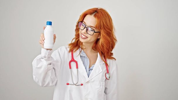 Junge rothaarige Ärztin lächelt zuversichtlich und hält Medikamentenflasche über isoliertem weißem Hintergrund - Foto, Bild