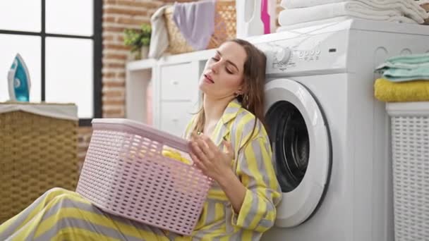 Mujer rubia joven sosteniendo cesta con ropa con expresión triste en la lavandería - Imágenes, Vídeo