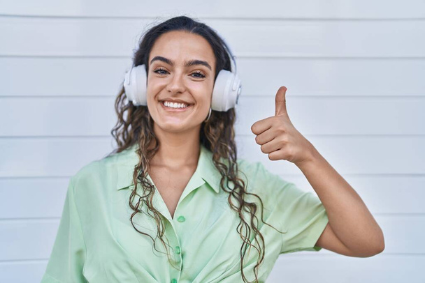 Młoda Latynoska słuchająca muzyki za pomocą słuchawek uśmiechnięta szczęśliwa i pozytywna, kciuk do góry robi doskonałe i znak aprobaty  - Zdjęcie, obraz