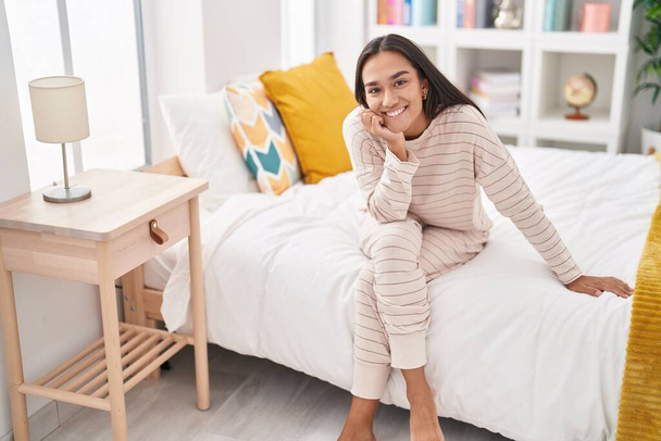 Νεαρή όμορφη ισπανόφωνη γυναίκα χαμογελά σίγουρος κάθεται στο κρεβάτι στο υπνοδωμάτιο - Φωτογραφία, εικόνα