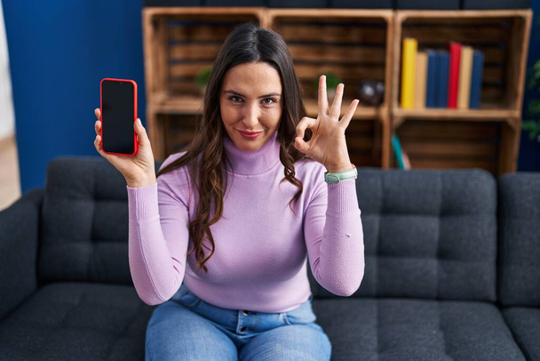 Молодая брюнетка, держащая в руках смартфон с пустым экраном, делает хорошо знак с пальцами, улыбаясь дружелюбный жест отличный символ  - Фото, изображение