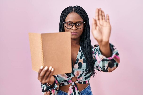 Afrikaanse vrouw met vlechten het lezen van een boek met open hand doen stop teken met ernstige en zelfverzekerde uitdrukking, verdediging gebaar  - Foto, afbeelding