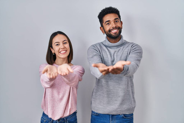 Mladý hispánský pár, který stojí vedle sebe a usmívá se dlaněmi, přijímá nebo gestikuluje. držení a ochrana  - Fotografie, Obrázek