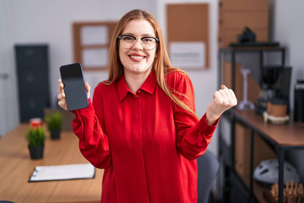 Punapää nainen työskentelee toimistossa osoittaa älypuhelimen näytön huutaa ylpeä, juhlii voittoa ja menestystä hyvin innoissaan nosti käsi  - Valokuva, kuva