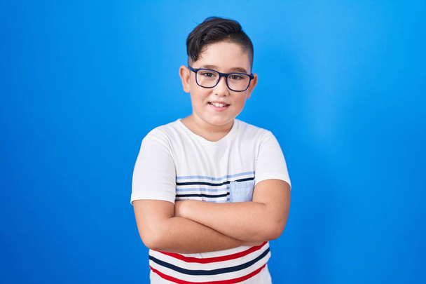 Jeune enfant hispanique debout sur fond bleu heureux visage souriant avec les bras croisés regardant la caméra. personne positive.  - Photo, image