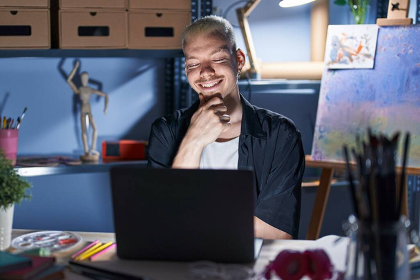 Young caucasian man using laptop at night at art studio looking confident at the camera smiling with crossed arms and hand raised on chin. thinking positive.  - Foto, imagen