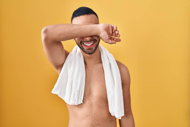Joven hombre hispano de pie sin camisa con toalla sonriendo alegre jugando a echar un vistazo a un boo con las manos mostrando la cara. sorprendido y salido  - Foto, Imagen