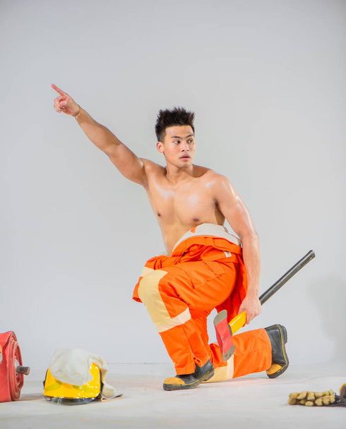 Topless firefighter is sitting and holding an iron axe while looking sideways and pointing a finger towards the top on a white background. - Photo, Image
