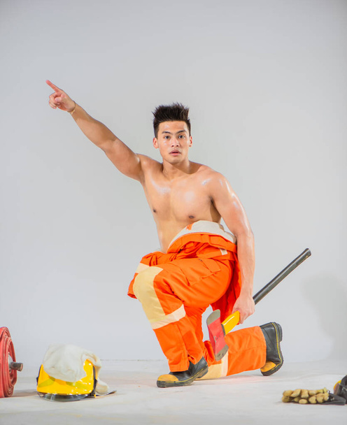 Topless firefighter is sitting holding an iron axe looking at the camera and pointing a finger towards the top. - Photo, Image