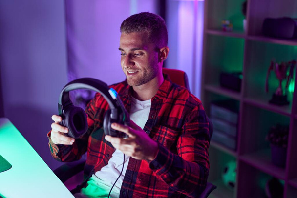 Молодий білий чоловік стример посміхається впевнено тримає навушники в ігровій кімнаті
 - Фото, зображення