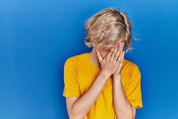 Молодой современный человек стоит на синем фоне с грустным лицом, прикрывающим лицо руками во время плача. депрессия.  - Фото, изображение