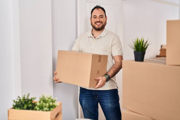 Jonge Spaanse man glimlachend zelfverzekerd met kartonnen pakket in nieuw huis - Foto, afbeelding