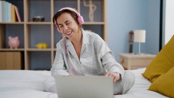 Латиноамериканка средних лет слушает музыку, сидящую на кровати в спальне - Фото, изображение