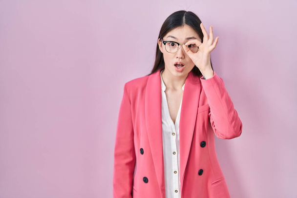Chinese business young woman wearing glasses doing ok gesture shocked with surprised face, eye looking through fingers. unbelieving expression.  - Photo, Image
