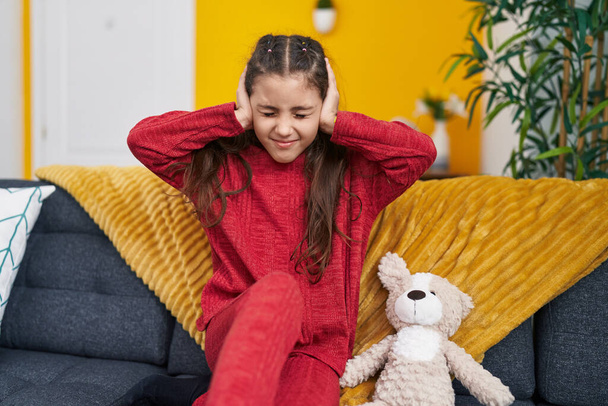 Adorable hispanic girl sitting on sofa covering ears for noise at home - Photo, Image