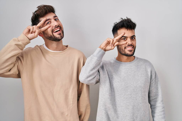 Young homosexual couple standing over white background doing peace symbol with fingers over face, smiling cheerful showing victory  - Fotoğraf, Görsel