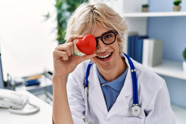 Νεαρός ξανθός άνδρας γιατρός χαμογελά σίγουρος κρατώντας την καρδιά πάνω από το μάτι στην κλινική - Φωτογραφία, εικόνα