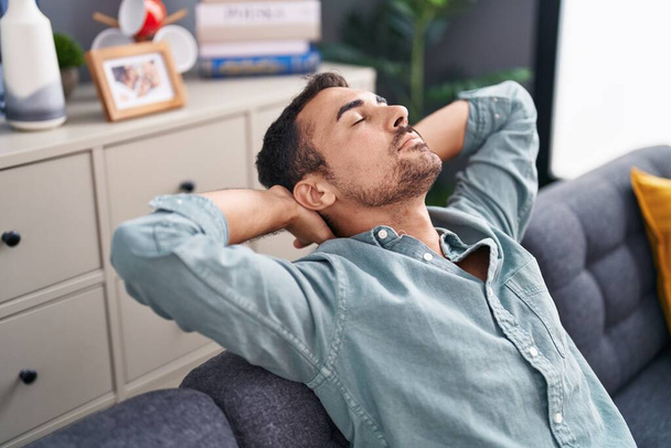 Jovem hispânico relaxado com as mãos na cabeça sentado no sofá em casa - Foto, Imagem