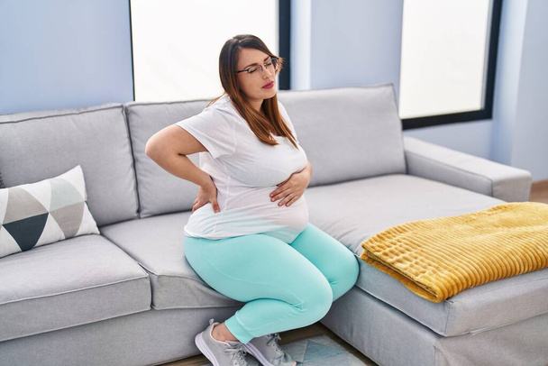 Νεαρή έγκυος γυναίκα που υποφέρει για οσφυαλγία κάθεται στον καναπέ στο σπίτι - Φωτογραφία, εικόνα