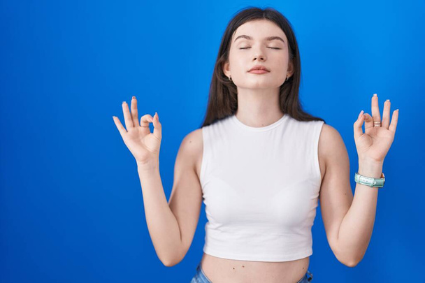 Mavi arka planda duran genç beyaz kadın rahatlamış ve parmaklarıyla meditasyon hareketi yaparken gözleri kapalı gülümsüyor. yoga konsepti.  - Fotoğraf, Görsel