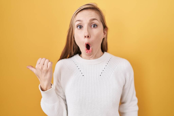 Mujer joven caucásica vistiendo suéter blanco sobre fondo amarillo sorprendido señalando con el dedo de la mano a un lado, boca abierta expresión asombrada.  - Foto, Imagen