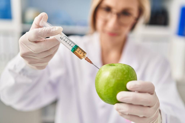Junge blonde Wissenschaftlerin spritzt im Labor Flüssigkeit auf Apfel - Foto, Bild