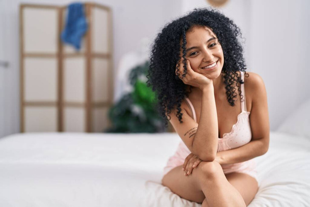 Молодая латиноамериканка улыбается уверенно сидя на кровати в спальне - Фото, изображение